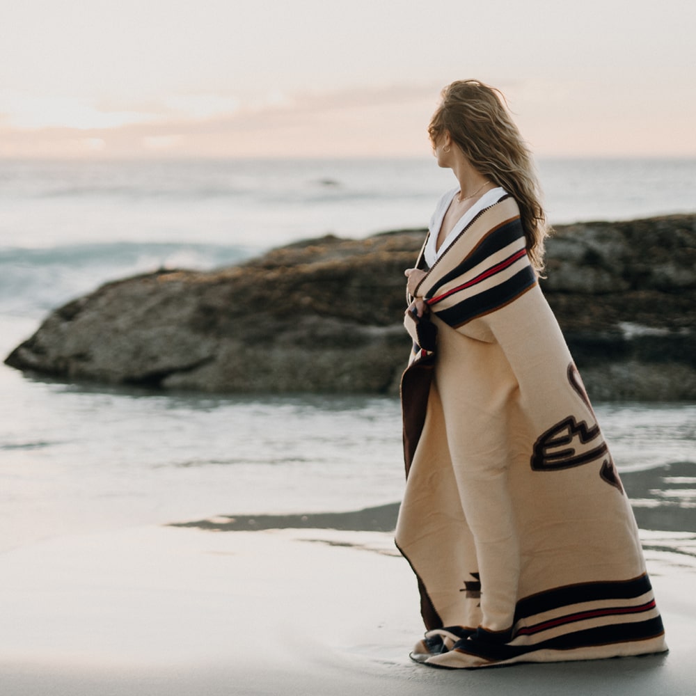 A woman wearing an Anifurry Thunderbird Merino Wool Blanket stands on the beach.